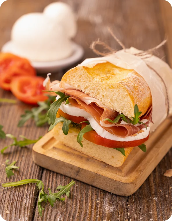 Italian-style sandwich on a rustic wooden board, filled with sliced cured meat, mozzarella, tomato, and arugula, with tomatoes and mozzarella blurred in the background.