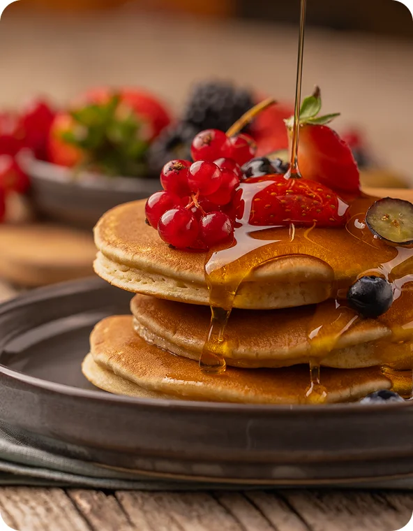 Stack of pancakes on a dark plate topped with strawberries, red currants, and blueberries as syrup is poured over, with a blurred bowl of berries in the background.