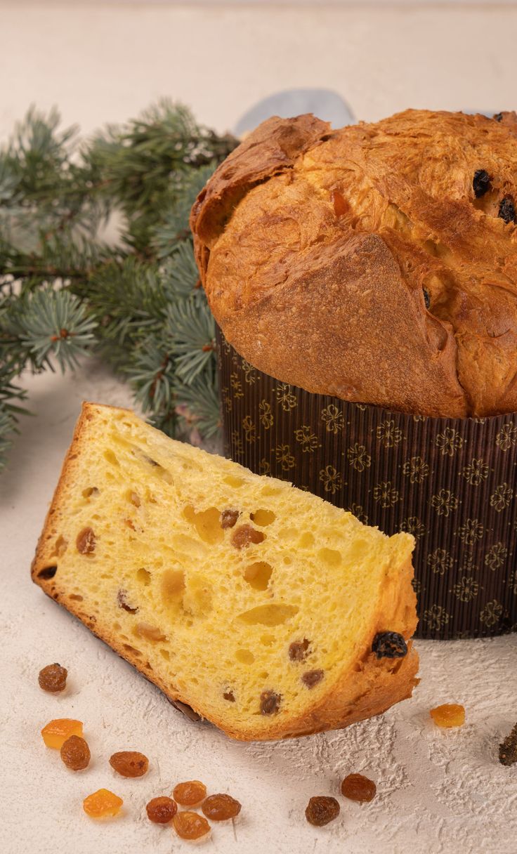 Panettone con fetta tagliata, uvetta e canditi, accanto a rami di abete su sfondo chiaro.