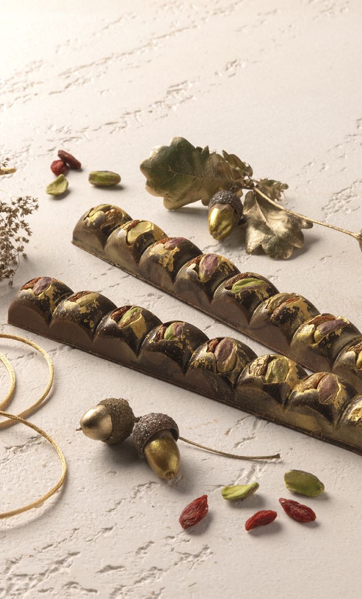 Due barrette di cioccolato con pistacchi e decorazioni dorate, tra ghiande e bacche su sfondo chiaro.