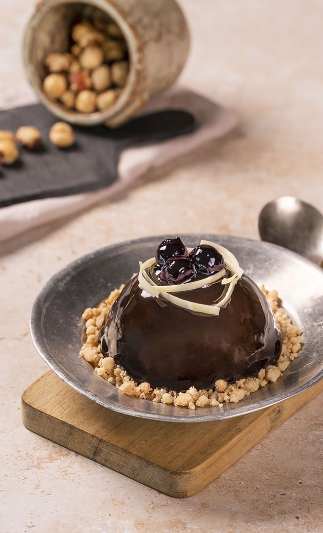 Cupola di cioccolato con frutti di bosco e riccioli di cioccolato bianco su crumble, in ciotola metallica.